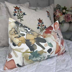 May include: Two decorative cushions with floral patterns. One cushion displays a watercolour-style design in shades of orange, green, and brown. The other has a cream background with embroidered red and blue floral sprigs. Both cushions are on a white quilted bedspread.