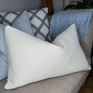 May include: A white throw pillow with a subtle herringbone pattern, placed on a beige couch with other pillows in shades of blue and white.