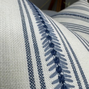 May include: A white and blue striped pillow with a decorative blue embroidered design. The pillow is made of a soft, textured fabric.