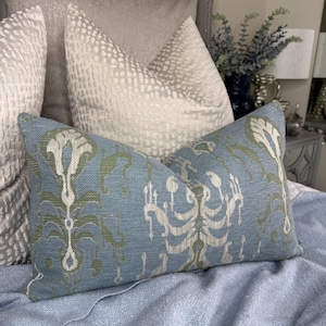 May include: A blue decorative cushion with a repeating pattern in green and off-white. The cushion is rectangular and rests on a light blue bedspread. Other pillows are visible in the background, creating a layered effect.