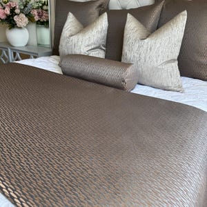 Op de afbeelding: Een bruin en beige bedsprei met een decoratief kussen en meerdere kussens. De bedsprei heeft een subtiele glans en een textuurpatroon.