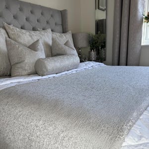 Puede incluir: Una cama con cabecero gris acolchado y almohadas a juego. Una colcha plateada-gris texturizada cubre la cama, con un edredón blanco debajo. La habitación tiene paredes y cortinas de tonos neutros.