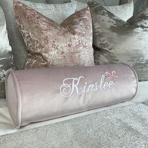 May include: A light pink velvet bolster pillow with the name "Kinslee" embroidered in white with a pink bow above the name.