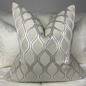 May include: A beige cushion with a silver geometric pattern. The cushion has a soft, plush appearance and a square shape. The geometric design is made up of silver lines on a beige background. This cushion would add a touch of elegance to any room.