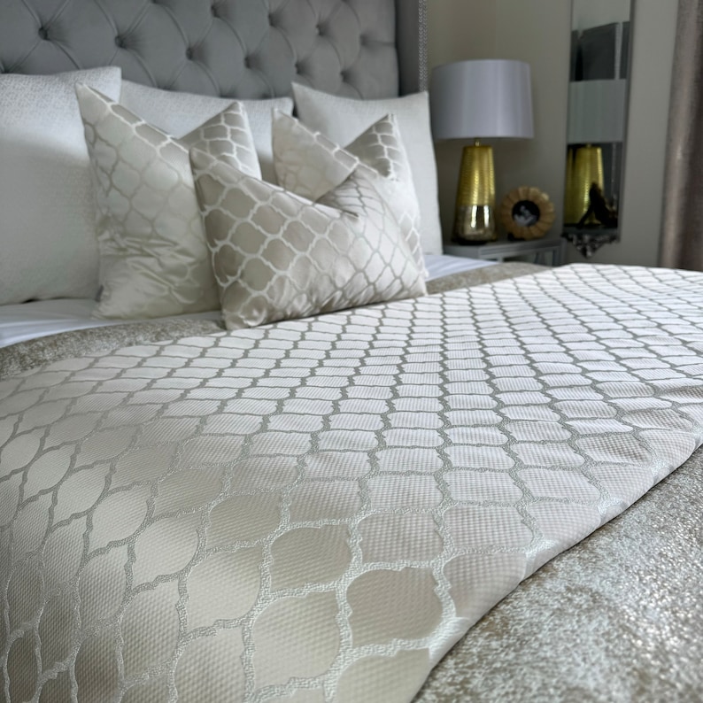 May include: A white bed with a white patterned bedspread and several white pillows. The bed is against a grey headboard with tufted buttons.