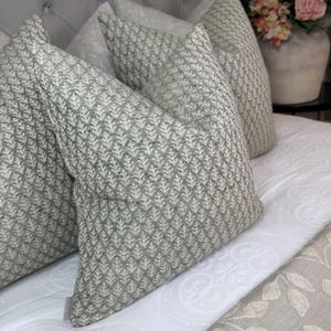 May include: Four decorative throw pillows with a light green and white leaf pattern. The pillows are arranged on a white bedspread with a subtle floral design. The pillows have a textured appearance and are in various sizes.