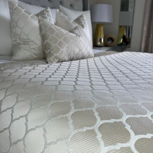 May include: A white bedspread with a geometric pattern. The bedspread is made of a soft, textured fabric. The bedspread is on a bed with white pillows.