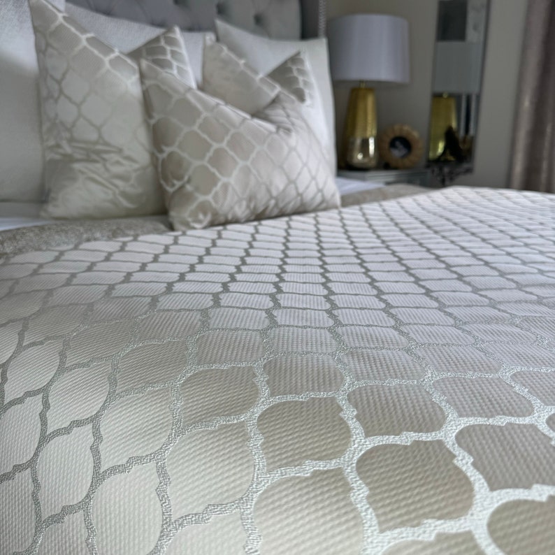 May include: A white bedspread with a geometric pattern. The bedspread is made of a soft, textured fabric. The bedspread is on a bed with white pillows.