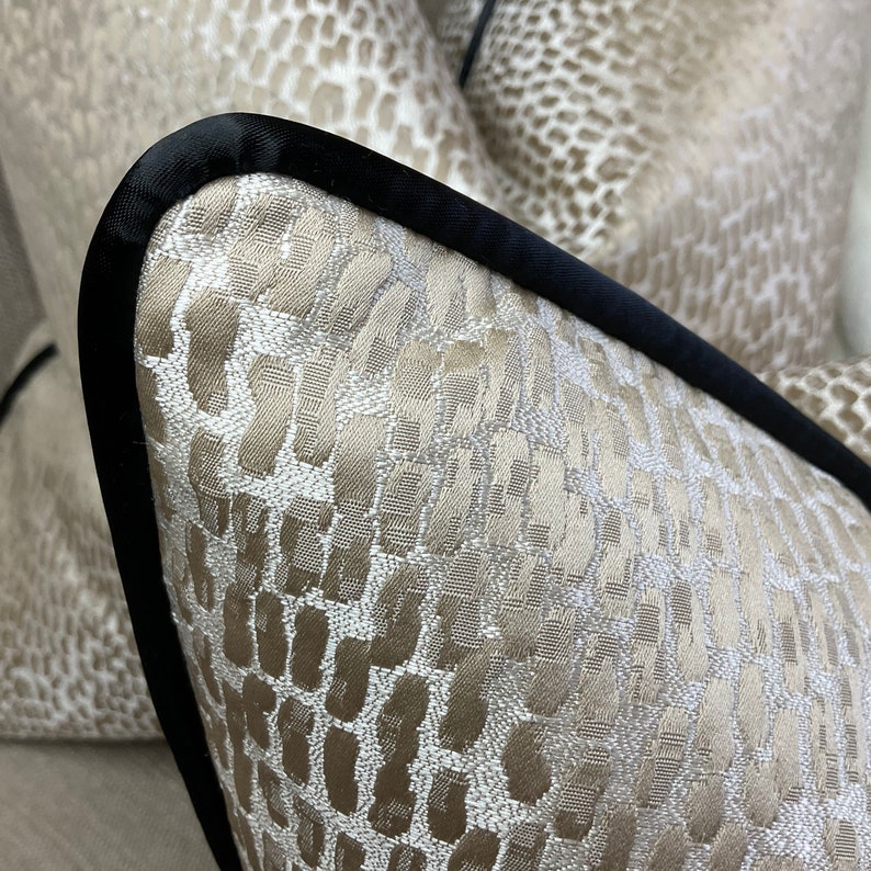 May include: Two beige throw pillows with a black trim. The pillows have a textured fabric with a subtle pattern.