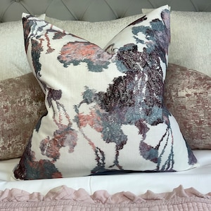 May include: A square decorative pillow with a white background and a floral pattern in shades of burgundy, gray, and peach. The pillow is placed on a bed with other pillows and bedding.