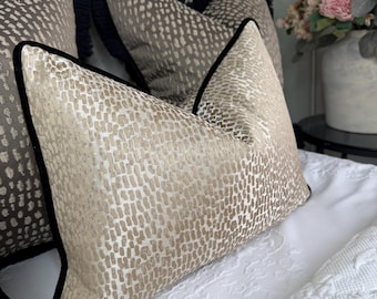 May include: Cushions with a beige dotted pattern and black trim. The cushions have a black fringe along the edges. The cushions are on a white bedspread.