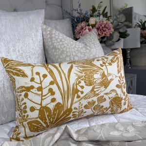 May include: A rectangular decorative pillow with a mustard-yellow bird and botanical print on a white background. The pillow is placed on a bed with other pillows and a floral arrangement in the background. The pillow measures approximately 12 x 20 inches.
