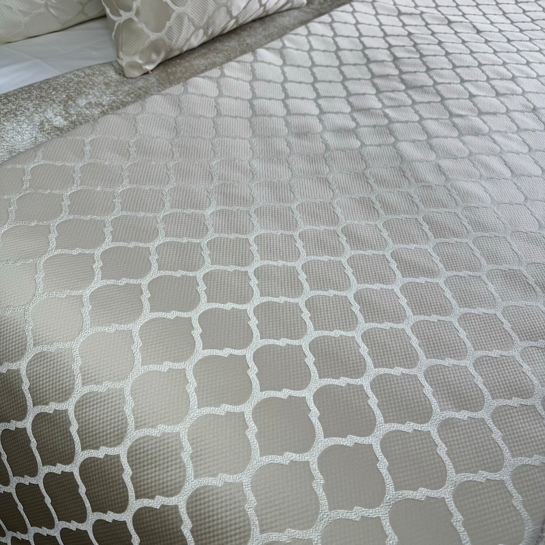 May include: A close-up of a white and beige patterned bedspread with a geometric design.