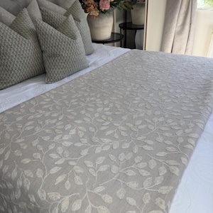 Puede incluir: Una cama con cabecero gris acolchado, varias almohadas grises estampadas y un camino de cama gris claro con un diseño de hojas bordadas. La cama está hecha con sábanas blancas.