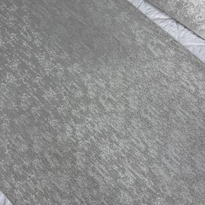 May include: A roll of textured gray wallpaper with a subtle, irregular pattern. The wallpaper has a matte finish and a slightly metallic sheen, creating a modern and elegant look. The design features vertical lines and small, scattered details.