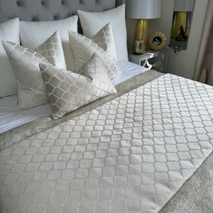 May include: A white bed with a white patterned bed runner and several white and cream patterned pillows. The bed is made with a white duvet cover.