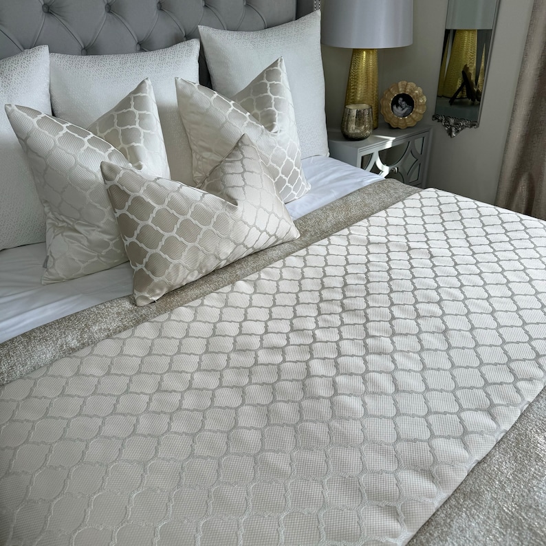 May include: A white bed with a white patterned bed runner and several white and cream patterned pillows. The bed is made with a white duvet cover.