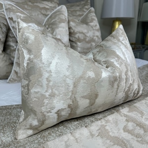 Puede incluir: Un conjunto de almohadas decorativas con un sutil patrón brillante de color gris claro y beige. Las almohadas están hechas de una tela suave y lujosa y tienen un brillo sutil.