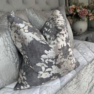 May include: Two decorative cushions with a floral pattern. The cushions feature a grey background with white and gold floral accents. The cushions are placed on a white quilted bedspread.