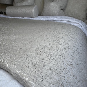 May include: A bed featuring a light grey textured comforter and matching pillows. The comforter has a subtle pattern, and the pillows include a bolster. The bedding is arranged on a white bedspread.