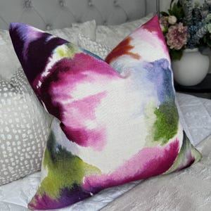 May include: A square throw pillow featuring a vibrant watercolor print with abstract splashes of magenta, deep purple, lime green, and soft blue on a white background. The fabric has a visible woven texture, creating a soft, artistic appearance.