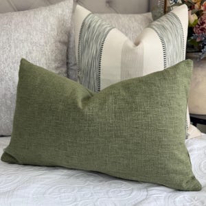 May include: A rectangular olive green throw pillow with a textured fabric finish, positioned in front of a square off-white pillow featuring vertical sage green striped patterns and a central cream panel, all resting on a white quilted bedspread.