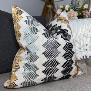 May include: A decorative throw pillow featuring a geometric pattern in shades of gold, black, and blue. The pillow is made from a soft, textured fabric with a subtle sheen.