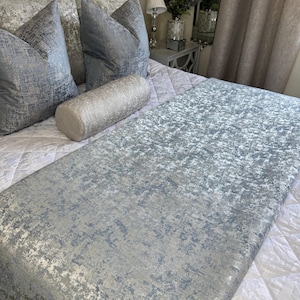 Op de afbeelding: Een bed met een grijs gestoffeerd hoofdbord, met een gewatteerde witte sprei en decoratieve kussens in zilver- en blauwtinten. Een zilveren bolsterkussen en een textuur bed runner maken de elegante slaapkamerinrichting compleet.