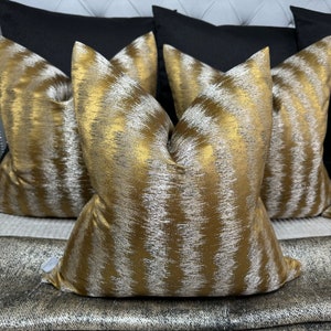 Puede incluir: Tres almohadas decorativas con un patrón geométrico dorado y plateado. Las almohadas están dispuestas en un sofá con estampado negro y plateado.