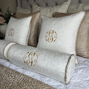 May include: A set of white and beige throw pillows with embroidered gold monograms. The pillows are arranged on a bed with a white and beige patterned bedspread.