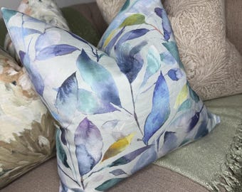 May include: Cushions with botanical leaf patterns in shades of blue, purple, and green. The cushions are on a neutral-toned sofa, with a green throw blanket visible. The cushions add a touch of nature-inspired decor.