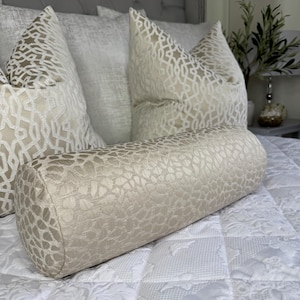 Puede incluir: Un cojín de apoyo beige con un patrón texturizado se encuentra sobre una cama blanca con almohadas decorativas estampadas.