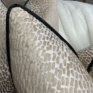 May include: Close-up of a beige pillow with a black trim and a subtle embossed pattern. The pillow is made of a soft, luxurious fabric.