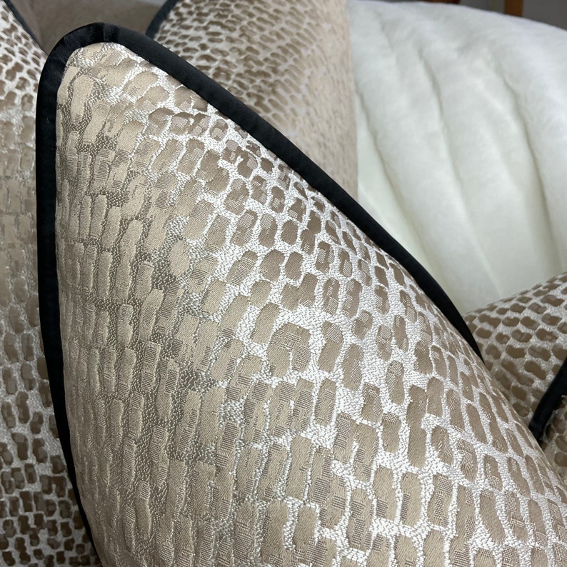 May include: Close-up of a beige pillow with a black trim and a subtle embossed pattern. The pillow is made of a soft, luxurious fabric.