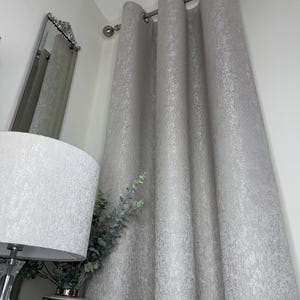 Puede incluir: Cortinas plateadas con ojales texturizados colgadas de una barra plateada. Las cortinas están junto a una lámpara blanca con una pantalla texturizada y un jarrón decorativo con vegetación. Se ve un espejo plateado.