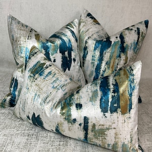 May include: Four decorative throw pillows with a white background and an abstract design in shades of blue, teal, and gold. The pillows have a soft, textured appearance, suggesting a comfortable and stylish home decor item.
