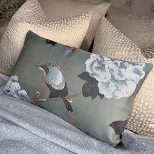 May include: A decorative rectangular pillow with a bird and floral design. The pillow features a teal background with a detailed embroidered bird perched on a branch, and large white and gray flowers. The pillow is surrounded by other decorative pillows.