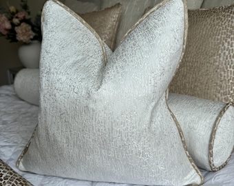 Op de afbeelding: Een wit kussen met een textuur en een beige rand. Het kussen ligt op een bed met andere kussens en een wit dekbed.