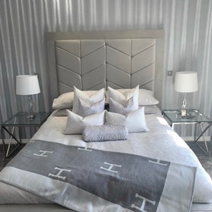 Pu&ograve; includere: Un letto imbottito grigio con una testata capitonn&eacute; e una coperta grigia e bianca a motivi. Il letto si trova in una camera da letto con carta da parati a righe grigie e due comodini con lampade.