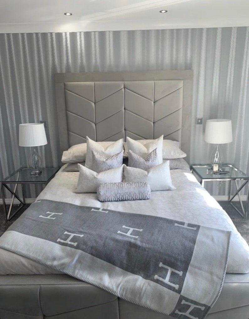 Pu&ograve; includere: Un letto imbottito grigio con una testata capitonn&eacute; e una coperta grigia e bianca a motivi. Il letto si trova in una camera da letto con carta da parati a righe grigie e due comodini con lampade.