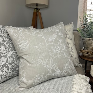May include: A set of three decorative throw pillows with a floral and bird design. The pillows are on a gray couch with a white throw blanket.