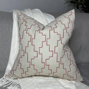 May include: A white cushion with a pink geometric pattern. The cushion is made of a soft, textured fabric and has a square shape.
