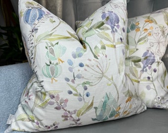May include: Two decorative throw pillows featuring a floral pattern. The pillows are white with a watercolour design of green leaves, purple flowers, and blue berries.
