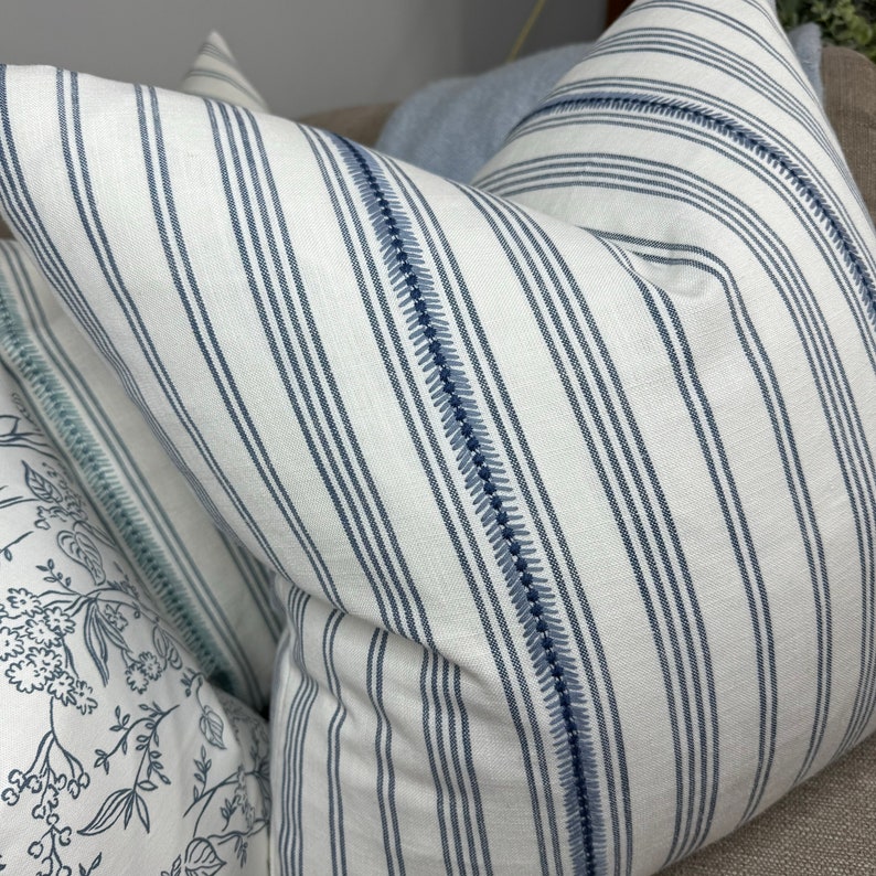 May include: A white and blue striped pillow with a blue stitched border. The pillow is made of a soft, textured fabric.