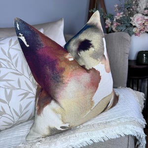 May include: A decorative cushion with a watercolour-style design featuring abstract shapes in shades of pink, orange, yellow, blue, and purple. The cushion is made of a soft, textured fabric and has a white background.