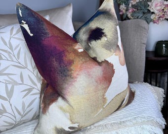 May include: A decorative throw pillow with a watercolor-style design featuring abstract shapes in shades of pink, orange, yellow, blue, and purple. The pillow is made of a soft, textured fabric and has a white background.