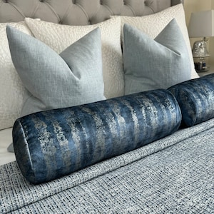 May include: Two light blue throw pillows with a white and grey patterned pillow behind them. A blue and silver patterned bolster pillow is in front of the pillows on a grey and white patterned bedspread.