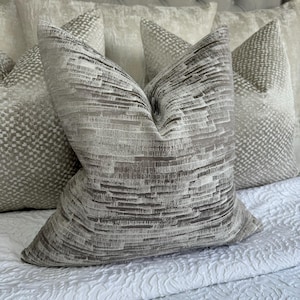 Pode incluir: Um conjunto de três almofadas decorativas com um padrão texturizado sutil em tons de cinza e branco. As almofadas estão dispostas sobre uma colcha branca.