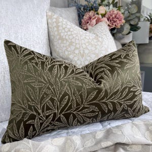 May include: An olive green cushion with a detailed leaf pattern. The rectangular cushion features a repeating design of leaves and vines in a lighter shade of green. It is placed on a bed with other cushions and floral arrangements.