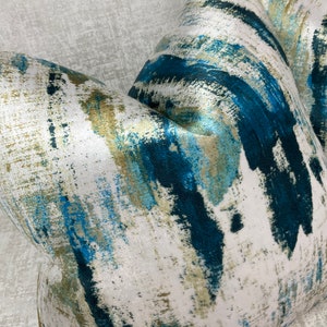 May include: Close-up of a decorative pillow with an abstract design. The pillow features a white background with brushstrokes of teal, blue, and gold. The fabric has a subtle sheen, adding to its luxurious appearance. The pillow is likely used for home decor.